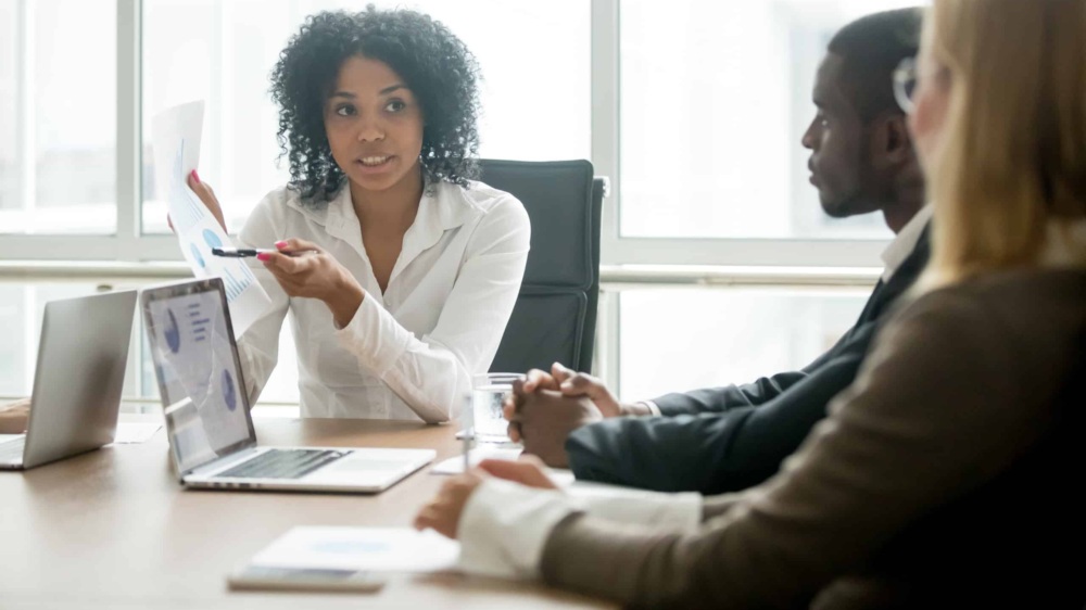 Black businesswoman showing a report on a piece of paper to a black man and white woman at a table in an office environment