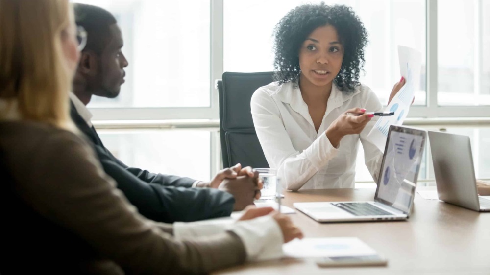 Black businesswoman showing a report on a piece of paper to a black man and white woman at a table in an office environment