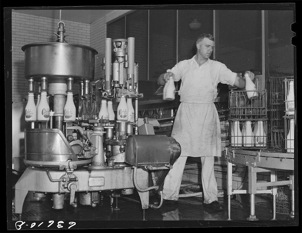 Black and white image of man taking milk bottles from milk capping machine and placing them in wire box crates for delivery