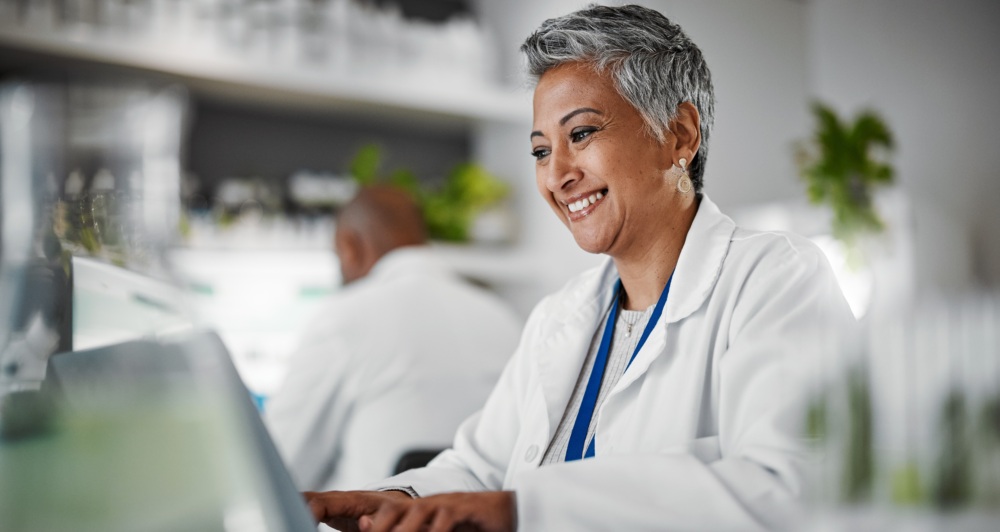 Middle-aged female scientist in white lab coat seated and typing on laptop in laboratory