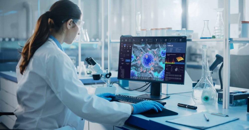 Woman in white lab coat and protective eyewear in a research lab at a computer with a virus on the screen