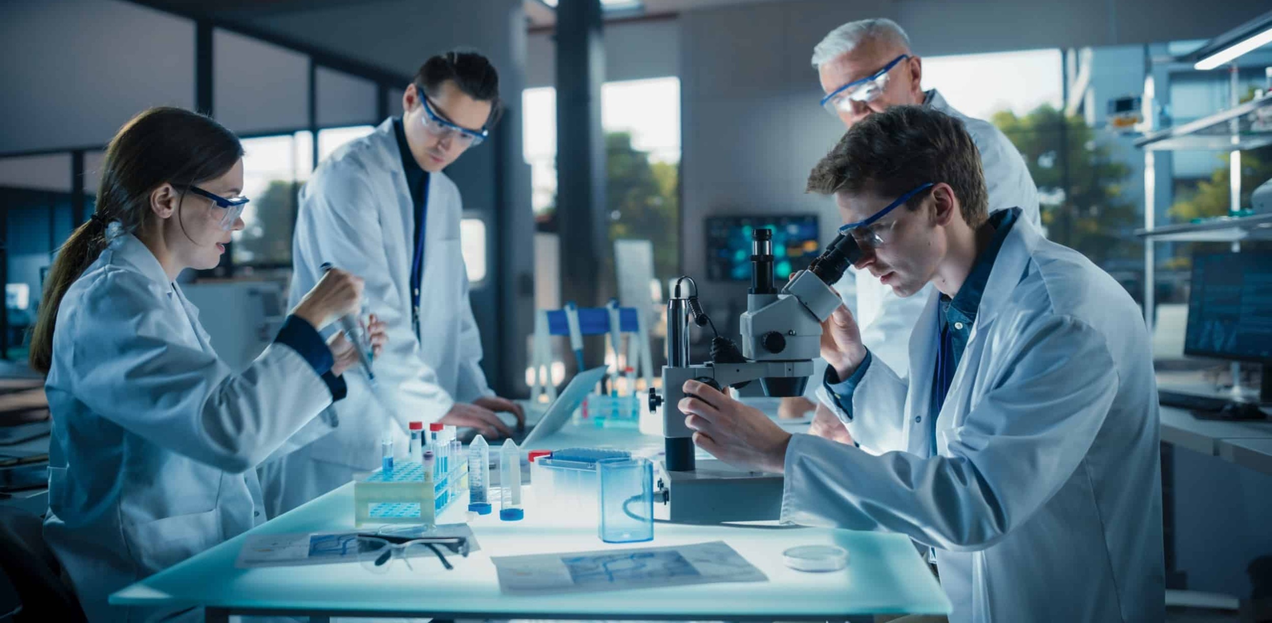 Four scientists in lab coats gathered around a lab table with a microscope, test tubes and other lab equipment